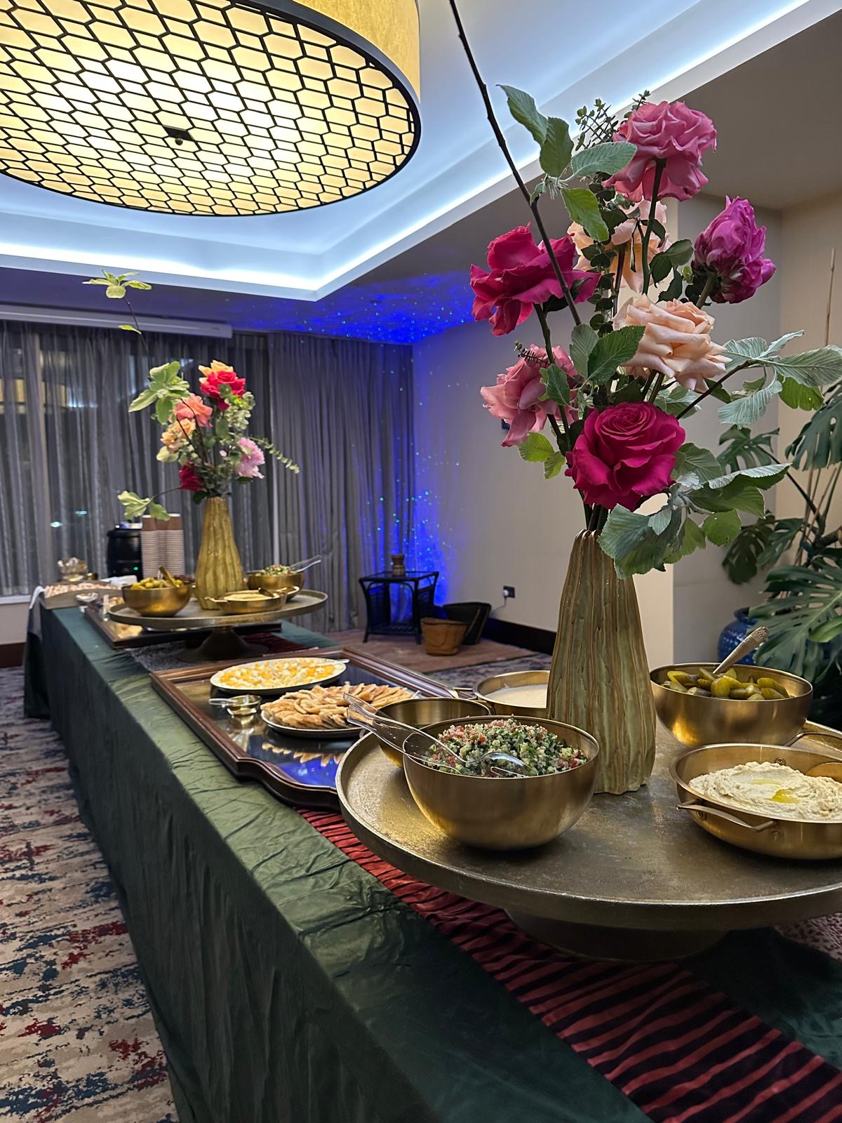 A long event buffet dressed in green with brass bowls of salads, dips and pickles, flatbreads and tall vases of pink and red roses.