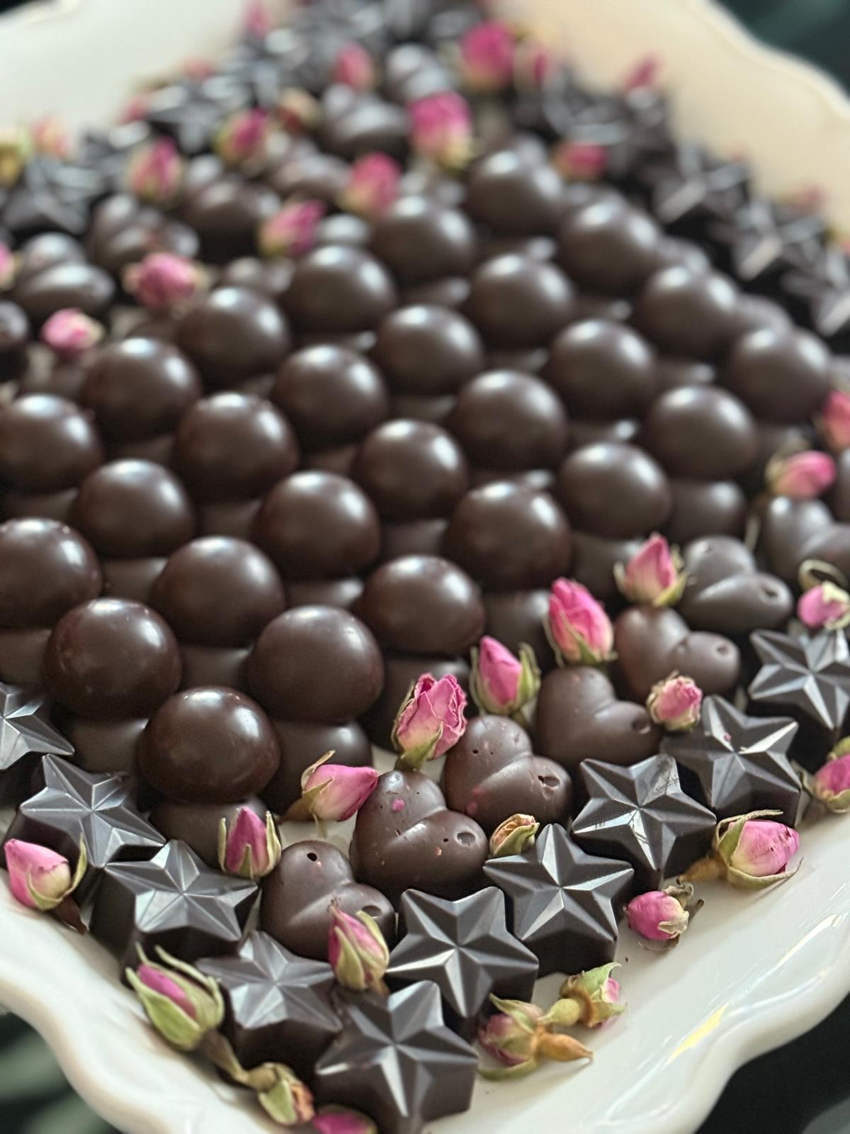 Close-up of glossy dark chocolate domes with chocolate stars and hearts and scattered dried pink rosebuds.