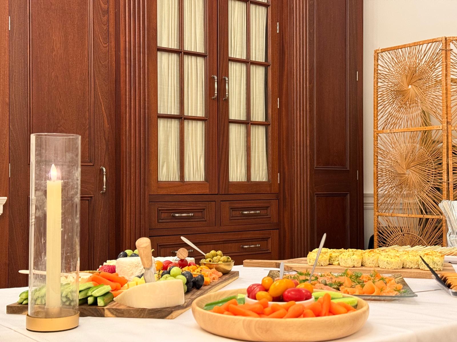 A white-clothed buffet with a cheese and fruit board, a wooden bowl of crudités, a platter of smoked salmon and a board of savoury bites, lit by a tall candle.